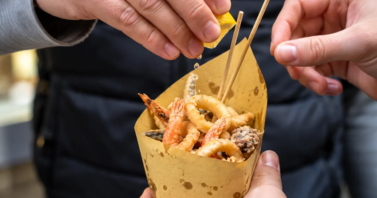 A hand holding a brown paper cone of fried seafood with a small wooden skewer, on a Naples street food tour
