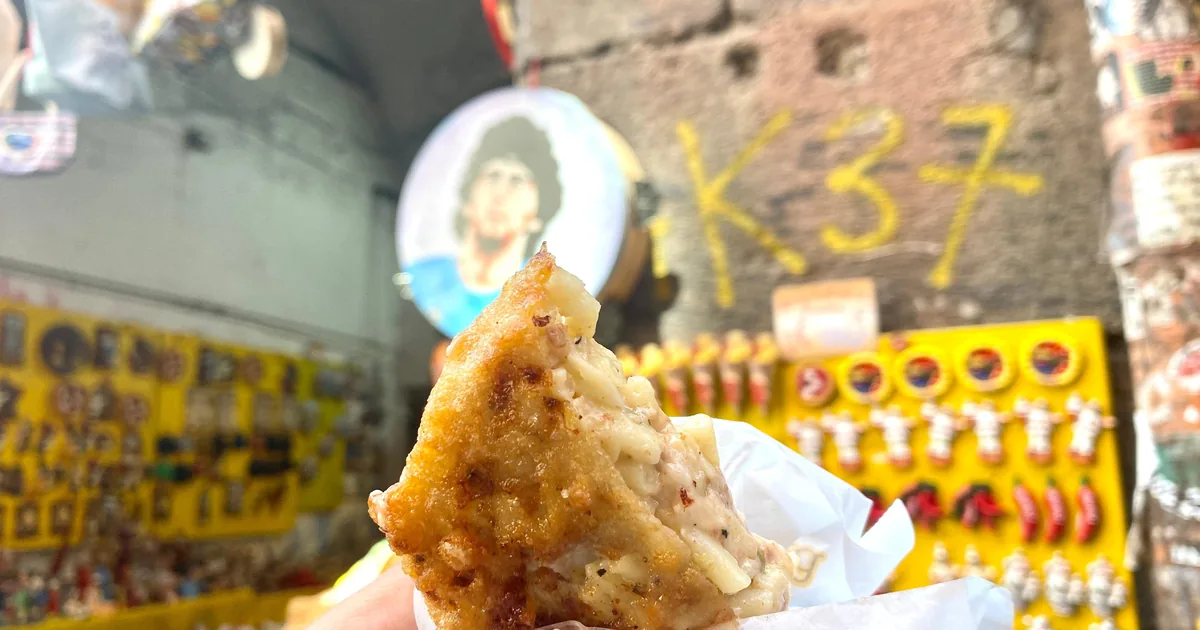 A hand holding a folded fried pizza in front of a street shrine to Maradona in the Quartieri Spagnoli